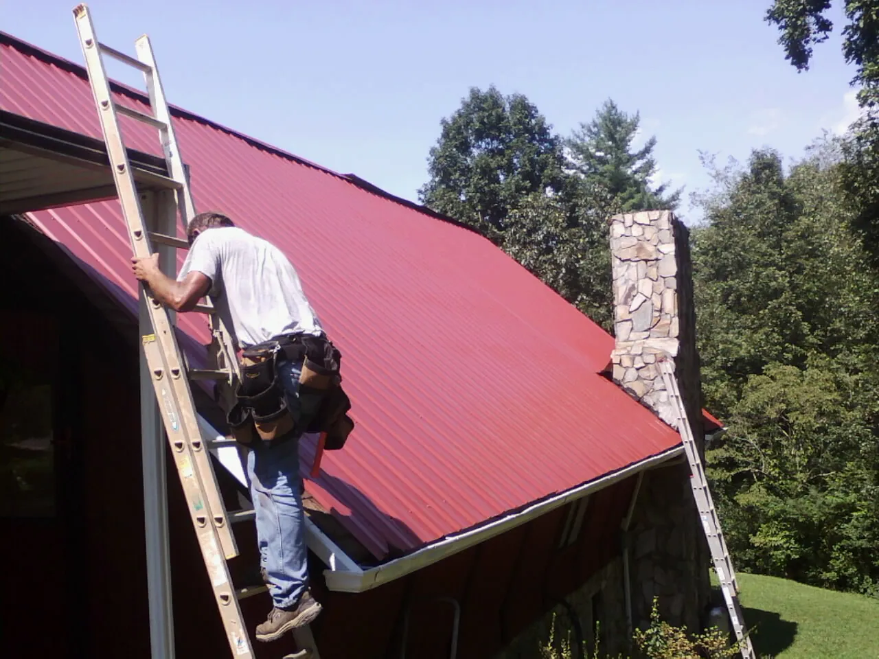 Roofer on a ladder beside a red metal roof and stone chimney — Soffit & Fascia Repair by Roofing Brothers in Taos